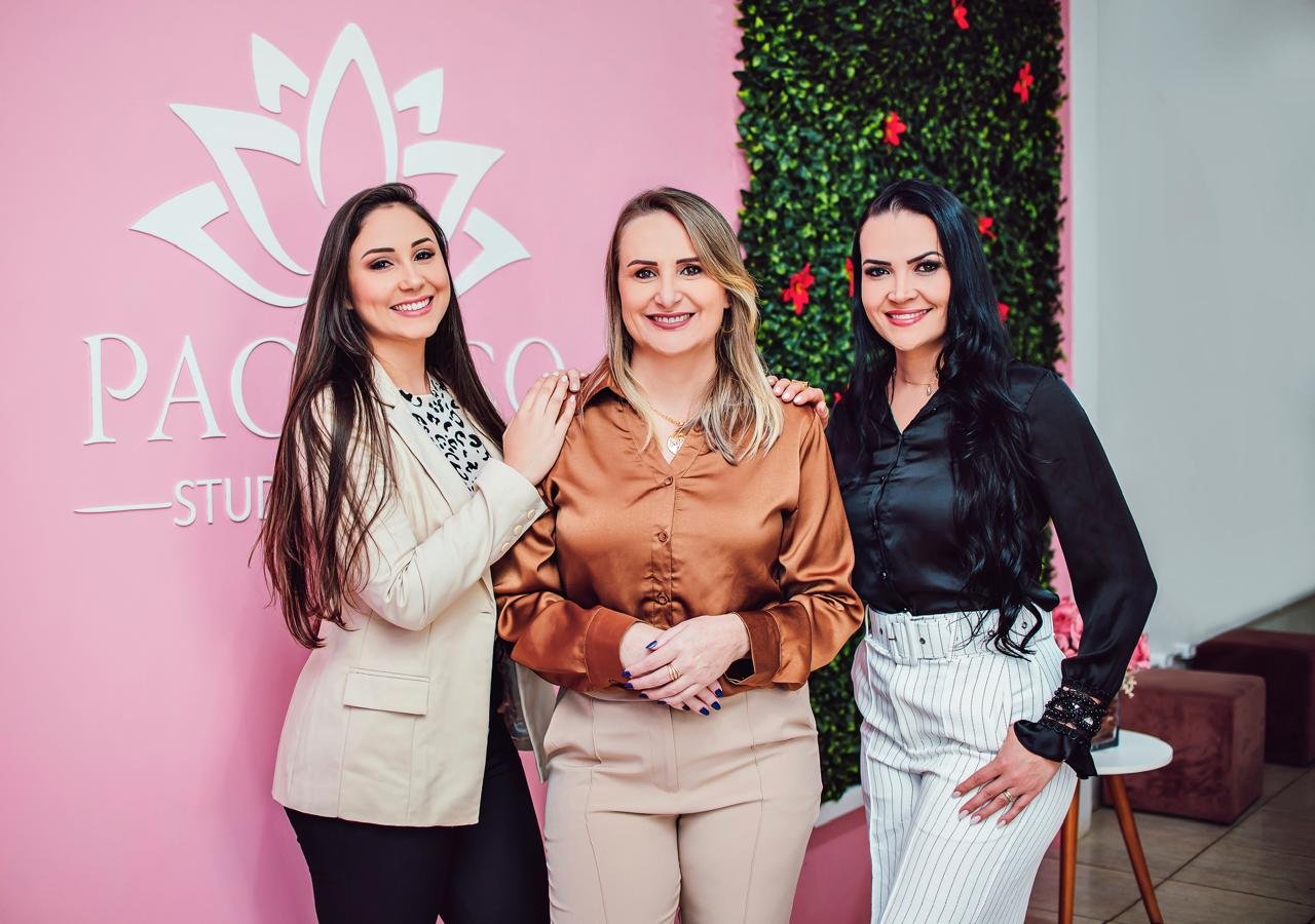 As três sócias do Pacheco Studio de Beleza lado a lado na recepção do espaço, com a logo de lótus na parede rosa